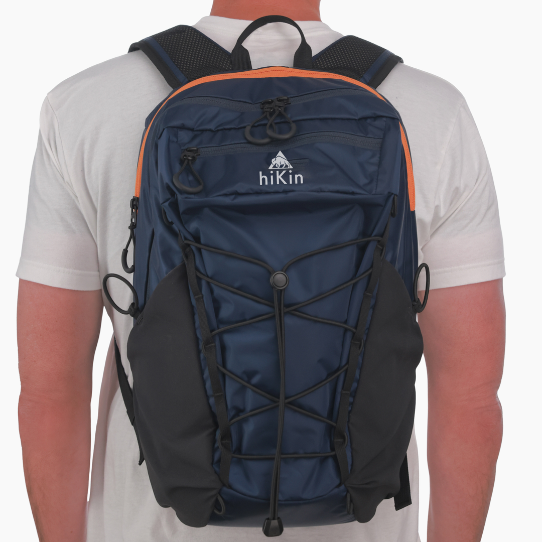 A person viewed from the back wearing a navy blue hikin backpack with black side pockets, black compression straps, and orange trim details.